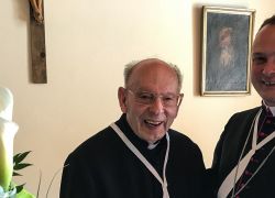 Zwei Männer in geistlicher Kleidung lächeln bei einer Feier im Stift Reichersberg neben Blumen und einem Kreuz.