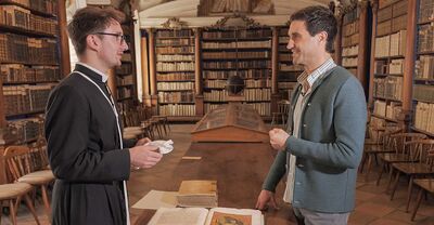 Ein Mann in traditioneller Kleidung diskutiert mit einem anderen vor einem Tisch in der Bibliothek des Stifts Reichersberg.