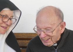 Eine Nonne und ein Priester lachen gemeinsam auf einer Bank beim Stift Reichersberg.