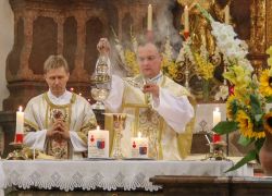 Ein Priester leitet ein feierliches Pontifikalamt im Stift Reichersberg, umgeben von Messdienern und blühenden Sonnenblumen.