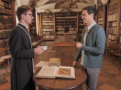 Zwei Männer unterhalten sich in der Bibliothek des Stift Reichersberg über ein geöffnetes Buch auf einem Holztisch.