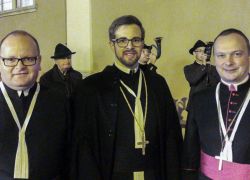 Drei Geistliche stehen bei der Abtbenediktion im Stift Reichersberg, während Männer im Hintergrund Hörner blasen.