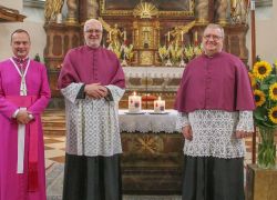 Drei Geistliche stehen lächelnd vor dem Altar von Stift Reichersberg, umgeben von Sonnenblumen und Kerzen.