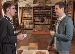 Ein Mann in traditioneller Kleidung diskutiert mit einem anderen vor einem Tisch in der Bibliothek des Stifts Reichersberg.