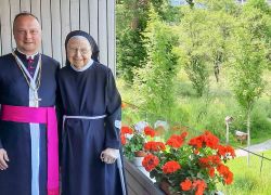 Ein Geistlicher und eine Nonne stehen lächelnd auf einem Balkon des Stift Reichersberg, umgeben von blühenden Geranien.