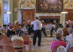 Ein Orchester spielt ein Abschlusskonzert im prächtig dekorierten Saal von Stift Reichersberg vor einem gespannten Publikum.