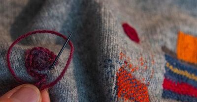 Eine Hand näht mit einer Nadel bunte Flicken auf einen grauen Stoff, inspiriert von der Kreativität im Stift Reichersberg.